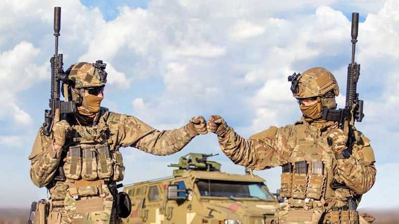 Двоє військових у формі і з автоматами дають один одному кулачок. Позаду видно джип